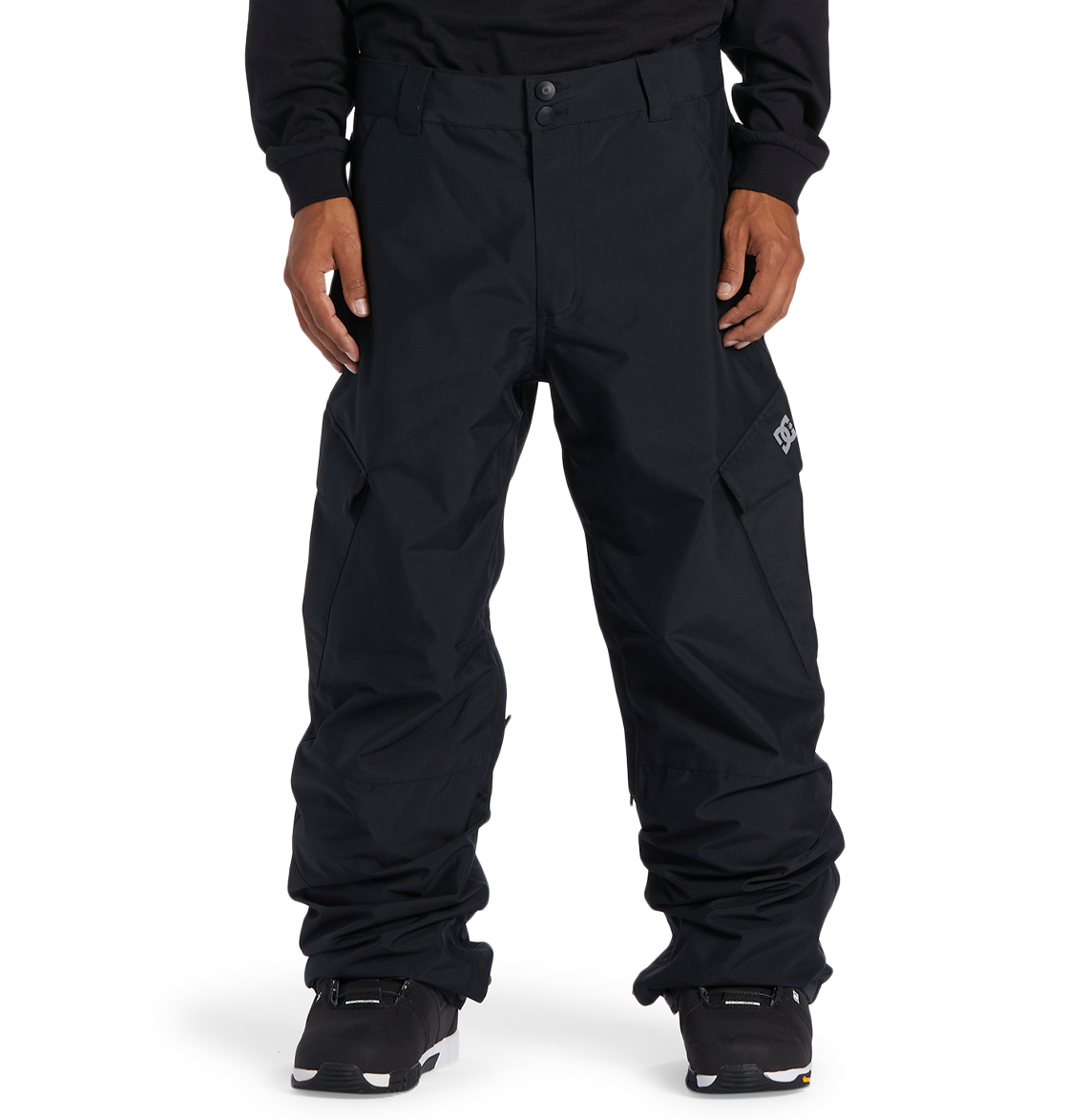 A person wearing loose-fitting black DC Shoes Men’s Banshee Insulated Pants, paired with a black sweatshirt and black sneakers, stands against a plain white background. The pants, designed as essential winter gear by DC Shoes, feature side pockets and a small white logo on the left thigh.