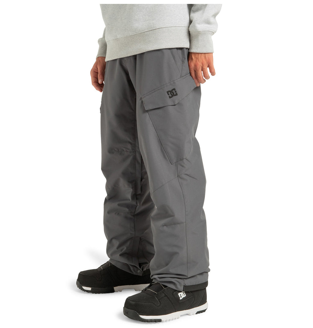 A person is shown from the mid-torso down against a white background, wearing the DC Shoes Men’s Banshee Insulated Pants in gray—crafted from recycled polyester and designed for winter mountain wear—with their hands relaxed at their sides. The pants feature practical side flap pockets and are paired with black sneakers.