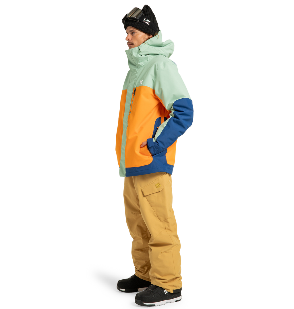 A person stands sideways with hands in pockets against a white background, wearing a mint, orange, and blue winter jacket; DC Shoes Men’s Banshee Insulated Pants crafted from recycled polyester in tan; black sneakers; a black beanie; and ski goggles.
