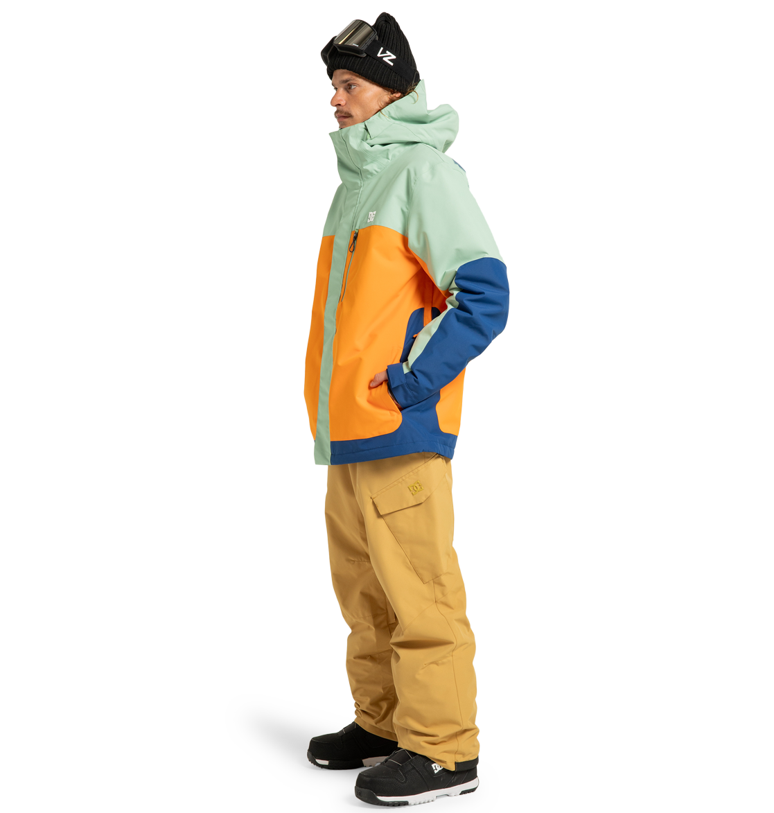 A person stands sideways with hands in pockets against a white background, wearing a mint, orange, and blue winter jacket; DC Shoes Men’s Banshee Insulated Pants crafted from recycled polyester in tan; black sneakers; a black beanie; and ski goggles.