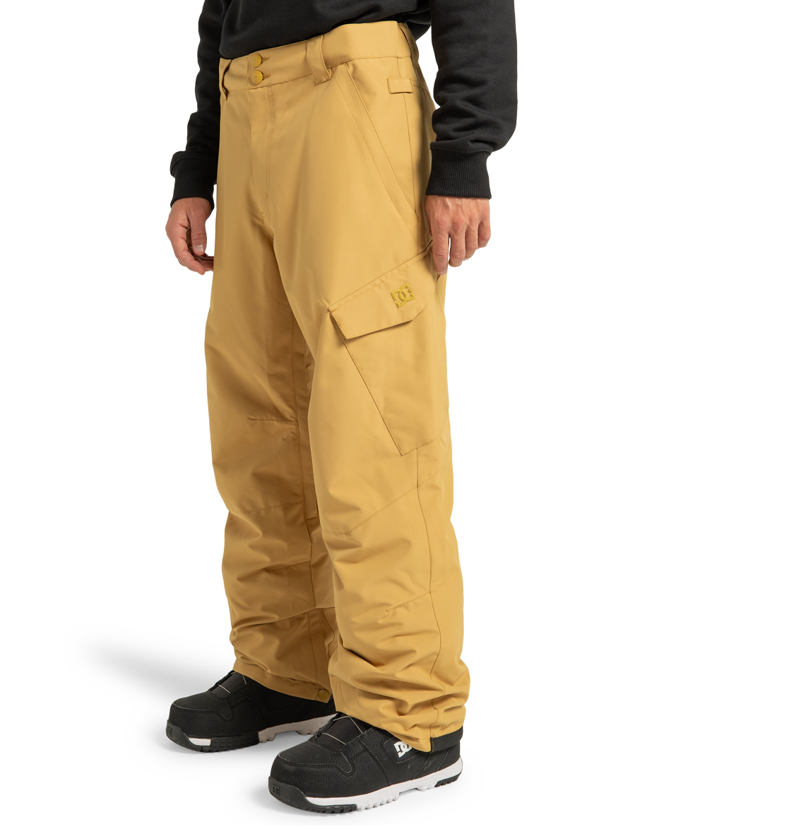 A person stands against a plain white background with their hands by their sides, wearing loose-fitting, tan DC Shoes Men’s Banshee Insulated Pants made from recycled polyester, paired with black sneakers and a black long-sleeve top.