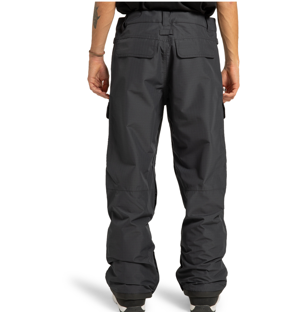 A person is standing with their back to the camera, wearing DC Shoes Men’s Code Shell Pants in dark grey and a black shirt. The pants have flap pockets, a loose fit, visible seams, and cuffed hems that fall over white shoes.