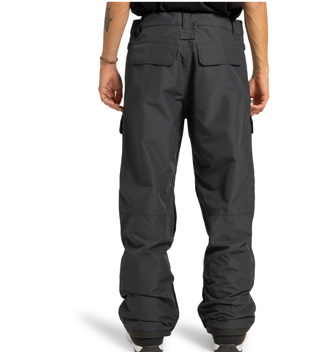 A person is standing with their back to the camera, wearing DC Shoes Men’s Code Shell Pants in dark grey and a black shirt. The pants have flap pockets, a loose fit, visible seams, and cuffed hems that fall over white shoes.