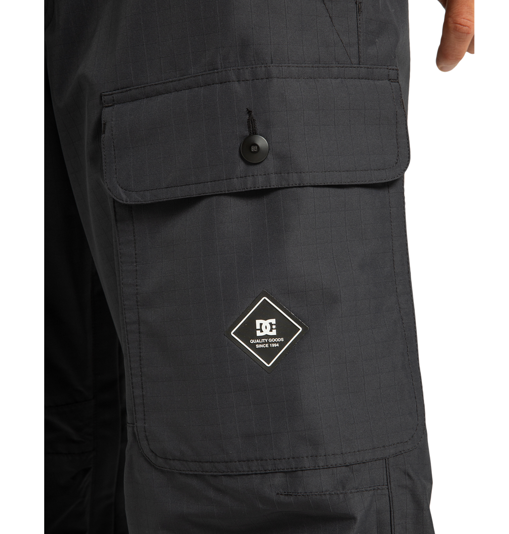 Close-up of a black cargo pocket on the DC Shoes Men’s Code Shell Pants, featuring a buttoned flap and a diamond-shaped patch with the DC logo and “Quality Goods” on the pocket.
