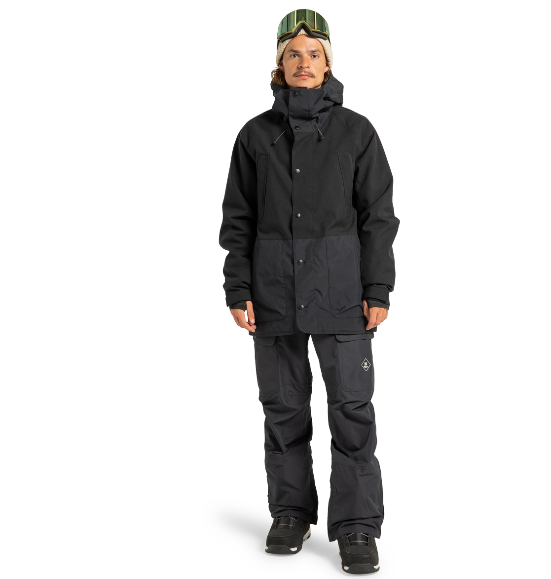 A person standing in snowboarding gear, featuring a black jacket, DC Shoes Men’s Code Shell Pants designed for eco-friendly performance, black boots, and green ski goggles resting on their head, against a plain white background.