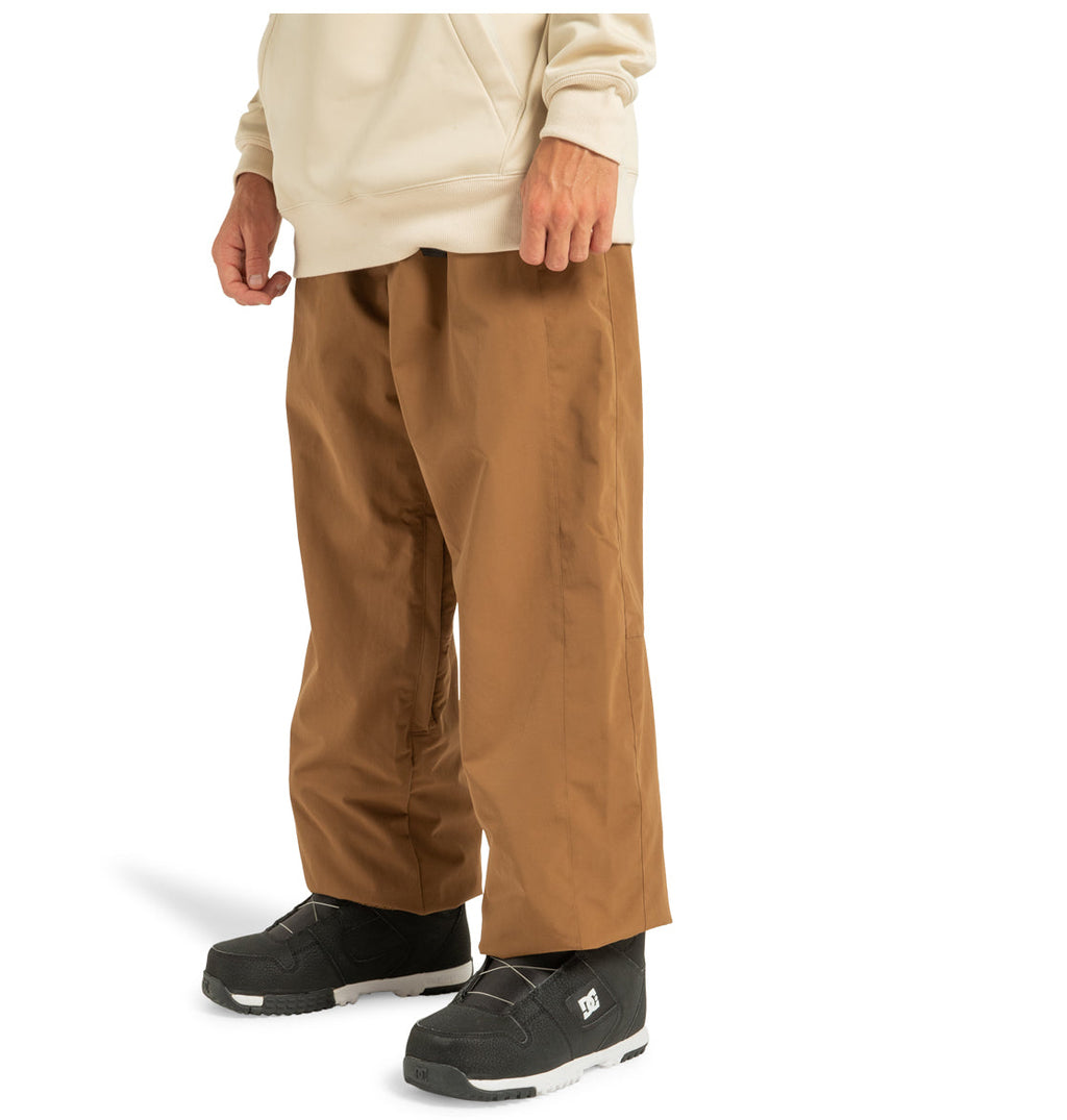 A person stands against a white background, showcasing only the lower half of their body and wearing a beige hoodie, black sneakers, and brown Unisex Primo Shell Pants by DC Shoes—renowned for weather defense and ideal for outdoor enthusiasts seeking both style and protection.