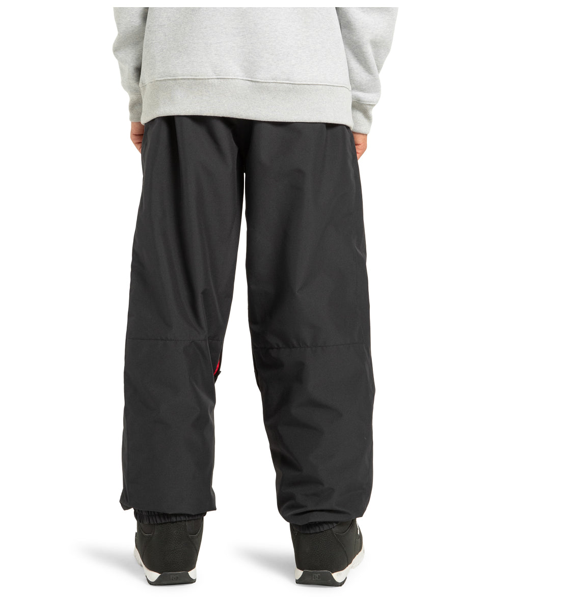 A person wearing DC Shoes’ Unisex Primo Snow Pants in black, paired with a grey sweatshirt and black-and-white sneakers, stands with their back to the camera. Only the lower half of their body is visible.