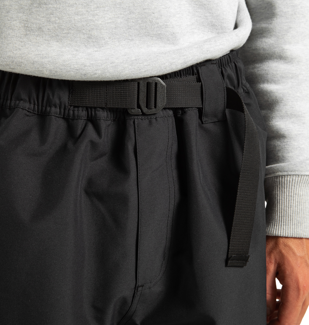 A close-up of a person wearing DC Shoes’ Unisex Primo Snow Pants in black, crafted from recycled polyester and featuring an adjustable webbing belt, paired with a gray sweatshirt. Only the lower torso and one hand are visible.