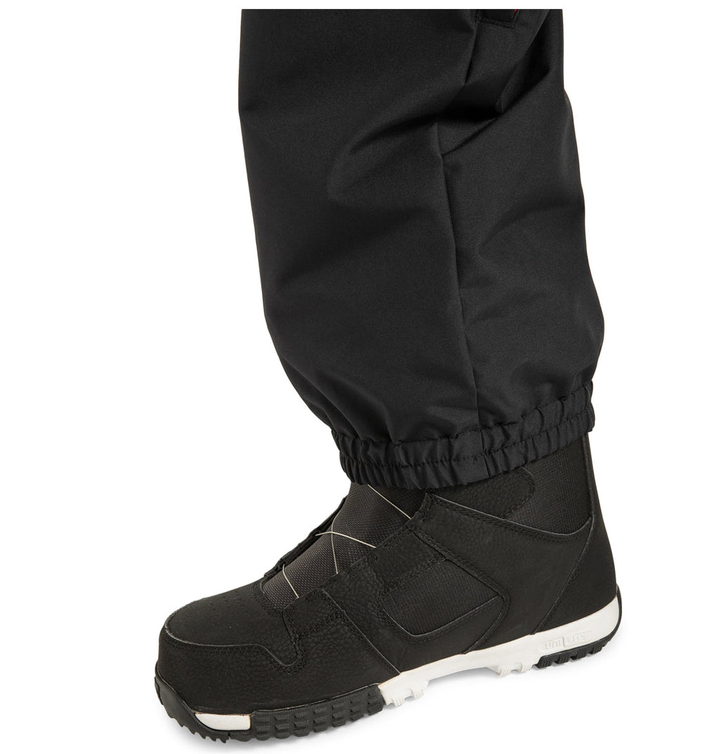 A close-up of a person wearing DC Shoes Unisex Primo Snow Pants, made from recycled polyester, paired with black snow boots featuring white soles, standing on a white background.