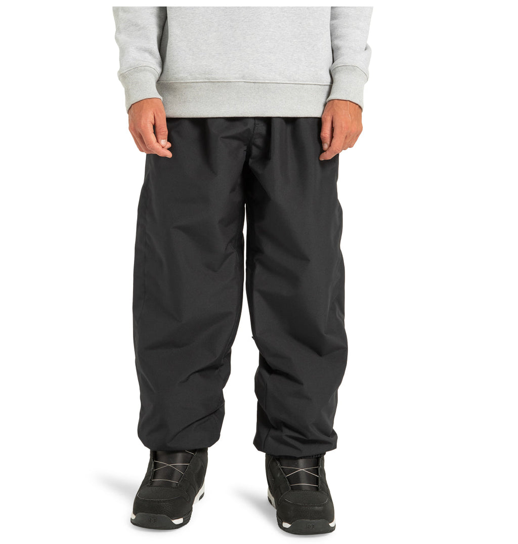 A person is wearing DC Shoes Unisex Primo Snow Pants in black, made from recycled polyester, along with a gray sweatshirt and black athletic shoes, standing against a plain white background.