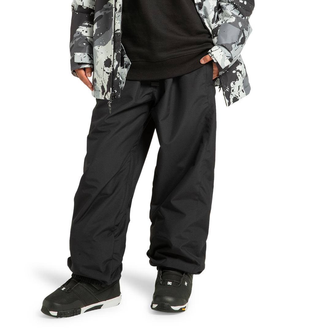 A person is shown from the waist down wearing DC Shoes Unisex Primo Snow Pants in black, paired with black sneakers, a black shirt, and a gray-and-black patterned jacket. The pants are made from recycled polyester, and the individual stands with hands in their pockets against a white background.