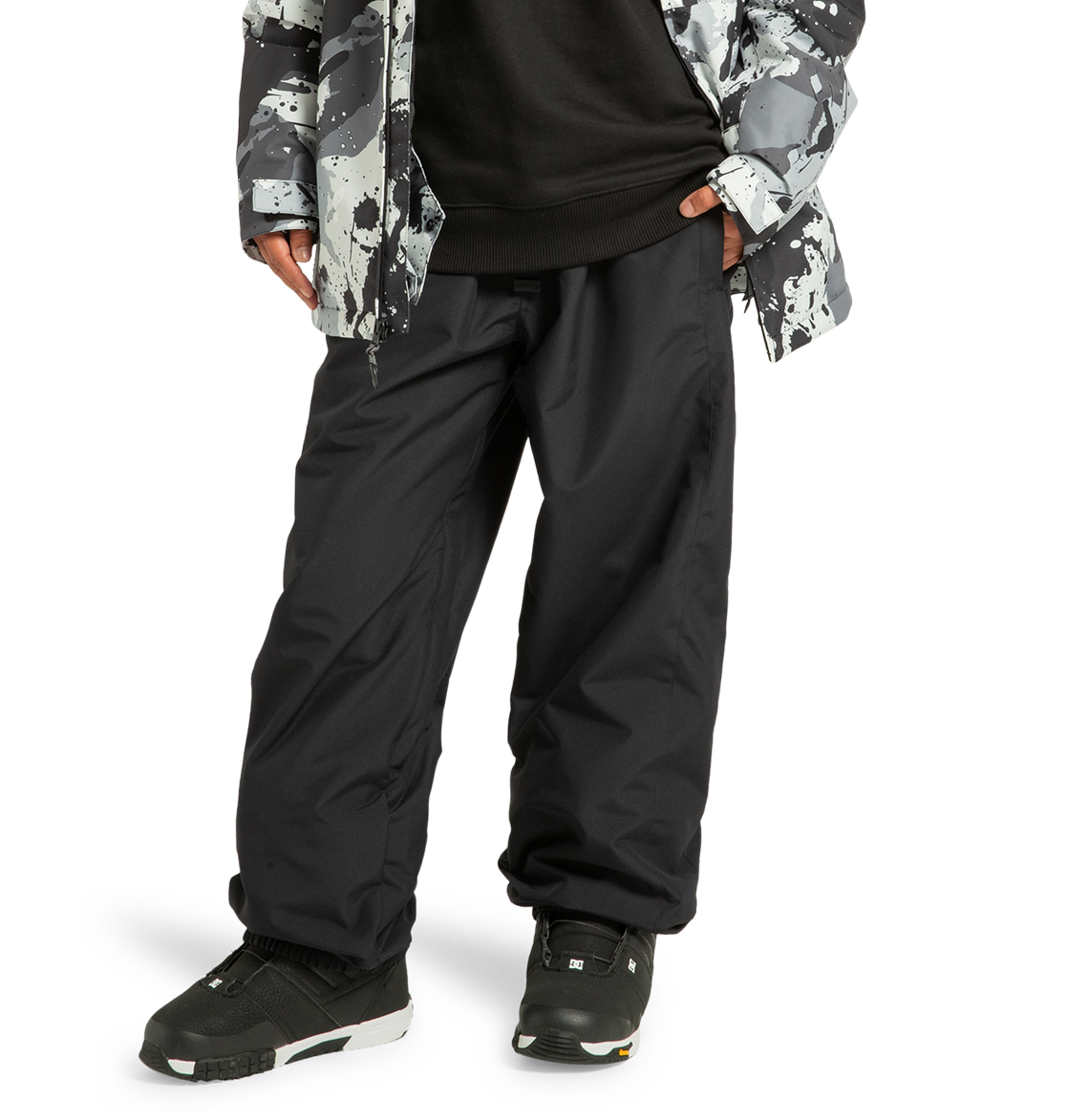 A person is shown from the waist down wearing DC Shoes Unisex Primo Snow Pants in black, paired with black sneakers, a black shirt, and a gray-and-black patterned jacket. The pants are made from recycled polyester, and the individual stands with hands in their pockets against a white background.