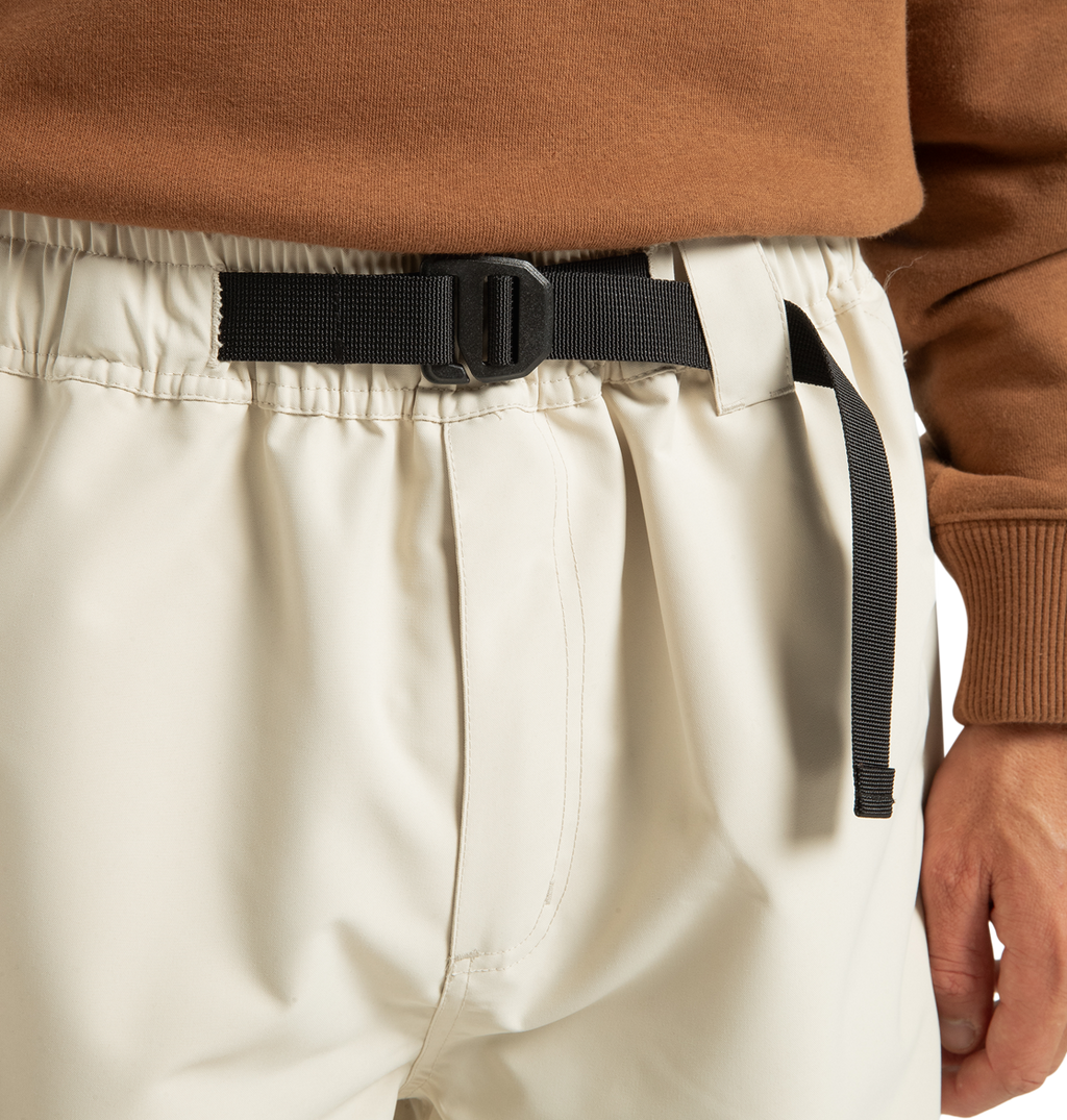 Wearing the Unisex Primo Snow Pants by DC Shoes in cream, featuring an elastic waistband with a black adjustable buckle belt and crafted from recycled polyester, styled with a brown long-sleeve top.