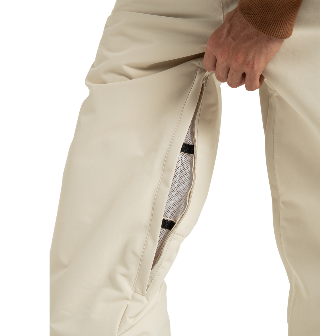 A person unzips a vent on the side of cream-colored DC Shoes Unisex Primo Snow Pants, revealing a mesh lining made from recycled polyester beneath. Only the lower torso and hand are visible.