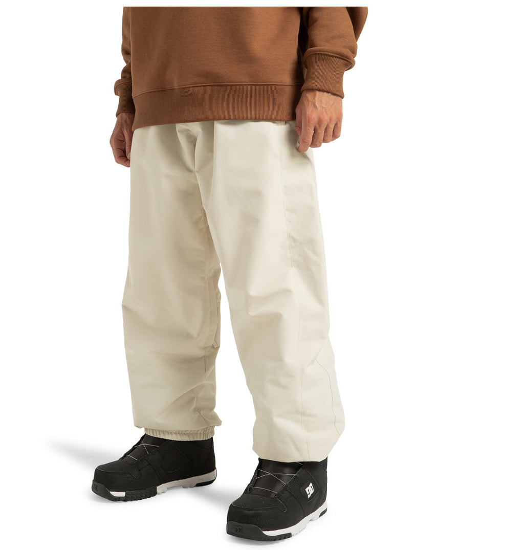 A person is shown from the waist down, wearing DC Shoes’ Unisex Primo Snow Pants—off-white, wide-leg snow pants crafted from recycled polyester—paired with a brown sweatshirt and black snow boots, all set against a white background.