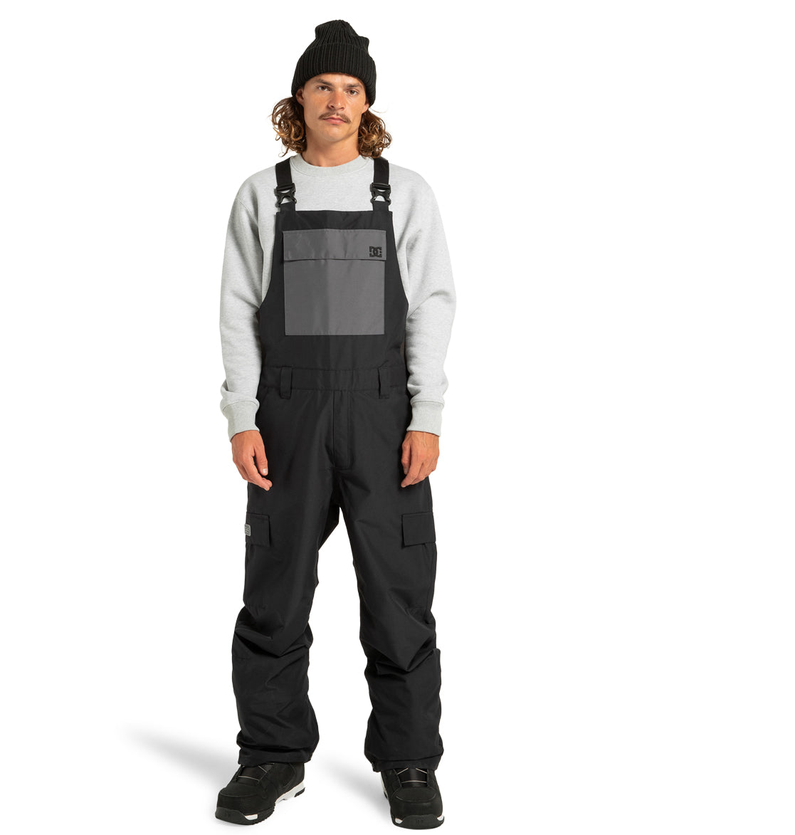 A person wearing a black beanie, gray sweatshirt, and DC Shoes Men’s Docile Bib Insulated Bib with a large front pocket stands facing forward against a white background.