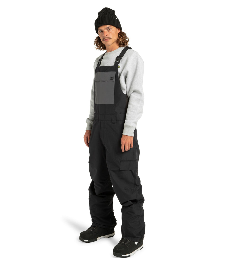 A man wearing a black beanie, gray sweatshirt, and the DC Shoes Men’s Docile Bib Insulated Bib with a gray chest pocket stands facing slightly left, hands relaxed at his sides, against a white background.
