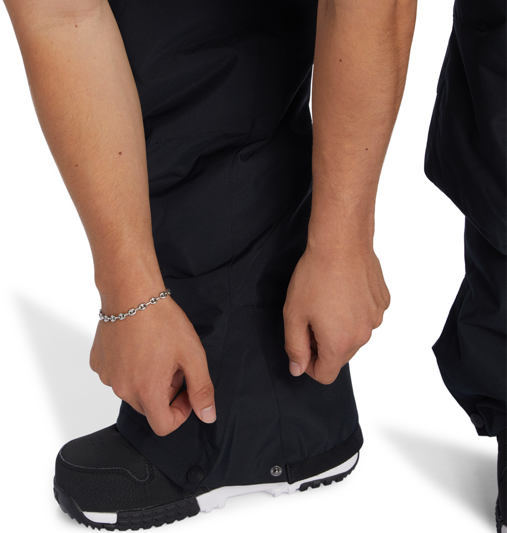 A person wearing DC Shoes Men’s Insulated Chino Pants in black and black-and-white shoes adjusts a snap at the ankle of their pants. Their left wrist has a silver chain bracelet, and the background is plain white.