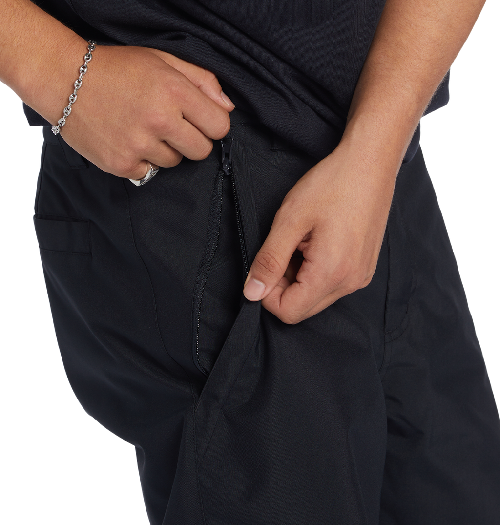A person, showing only their lower torso and hands, is unzipping a side pocket while wearing DC Shoes Men’s Insulated Chino Pants and a black shirt.