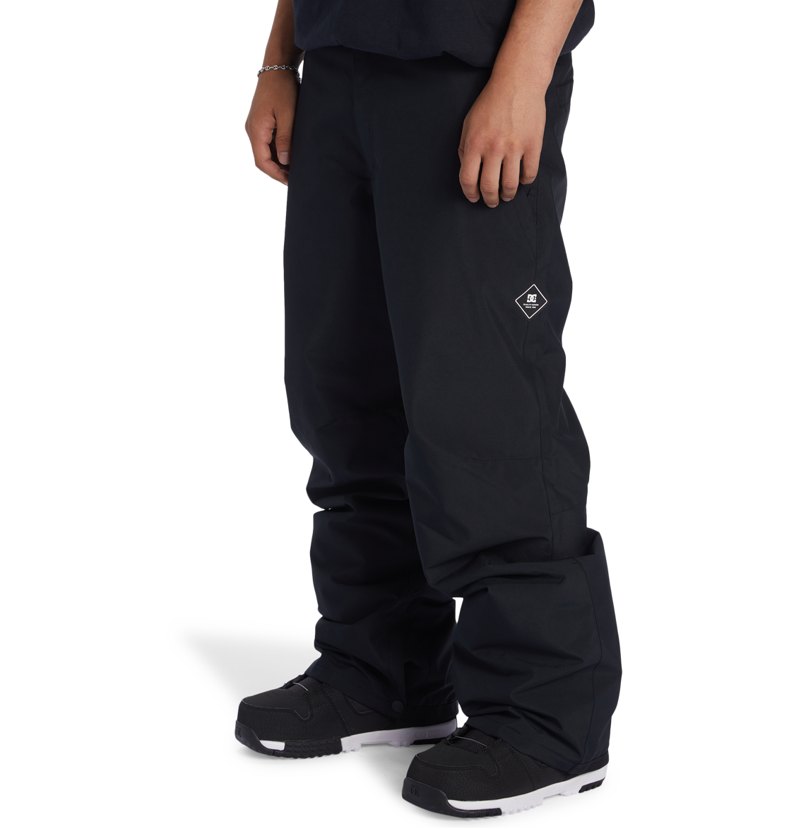A person is shown from the waist down wearing DC Shoes Men’s Insulated Chino Pants in black, featuring a small white DC Shoes logo on the left thigh, paired with black sneakers with white soles and standing against a plain white background.