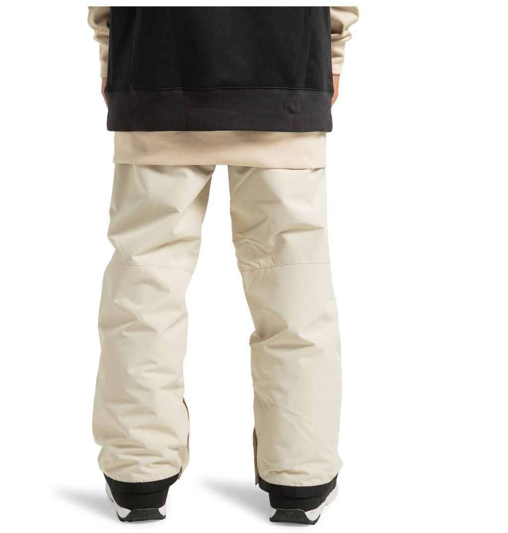A person wearing DC Shoes Men’s Insulated Chino Pants in a loose, light beige style made from recycled polyester, paired with a black and beige jacket. Only the lower half of their body, from mid-torso down, is visible from the back. They are also wearing black weather-defending shoes.