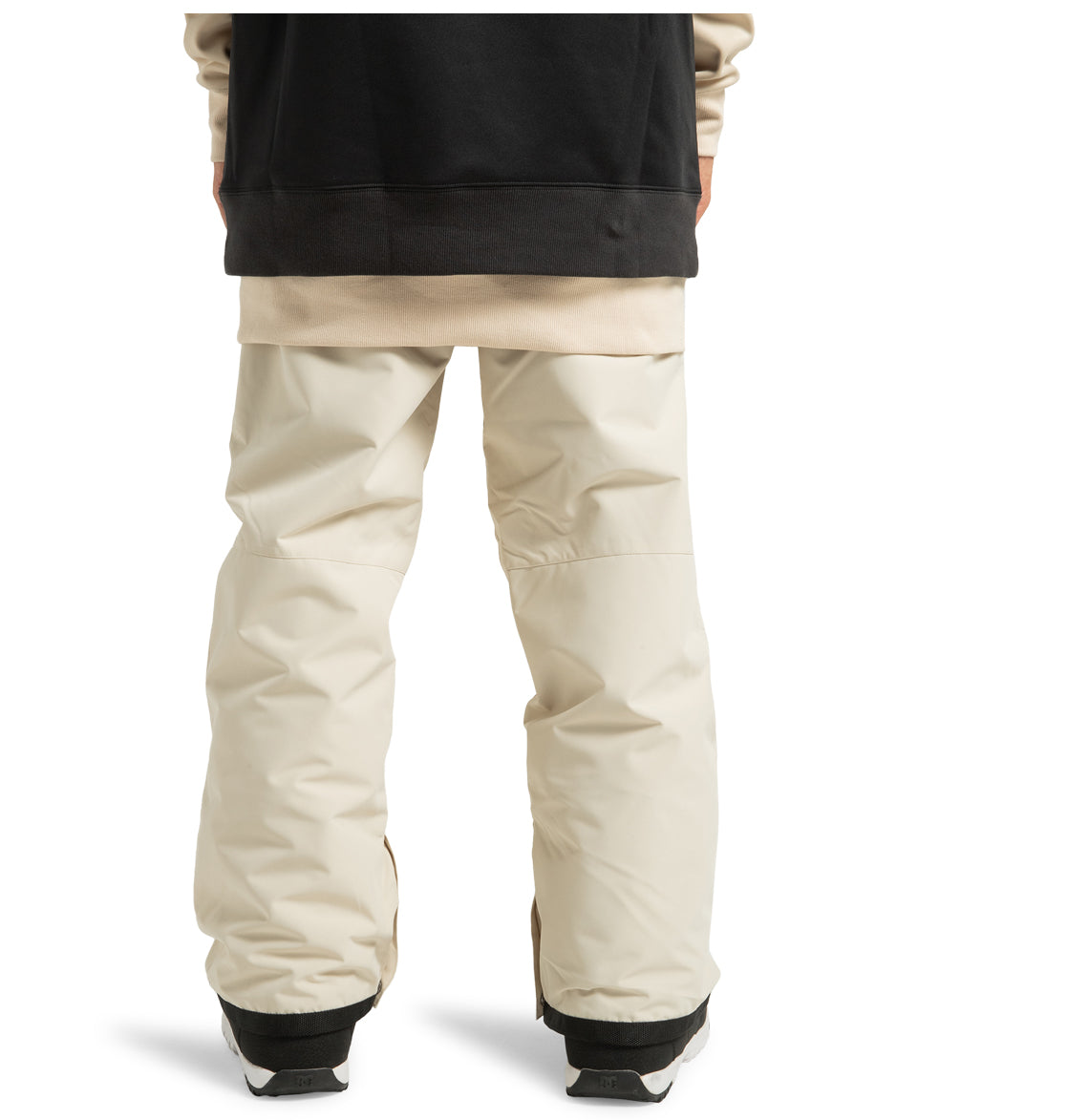 A person wearing DC Shoes Men’s Insulated Chino Pants in a loose, light beige style made from recycled polyester, paired with a black and beige jacket. Only the lower half of their body, from mid-torso down, is visible from the back. They are also wearing black weather-defending shoes.