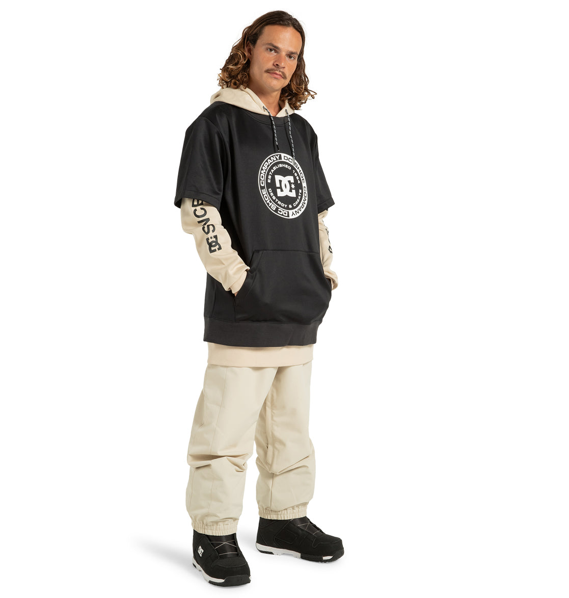 A man with long hair is wearing a two-tone beige and black DC Shoes hoodie, paired with DC Shoes Men’s Insulated Chino Pants crafted from recycled polyester in a matching beige color. He completes the look with black sneakers and stands with his hands in his hoodie pockets.