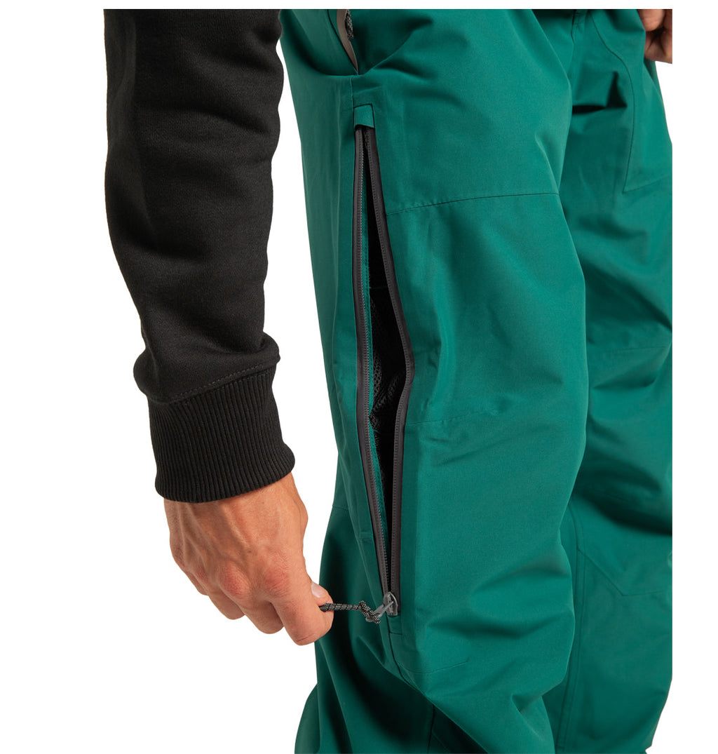 Wearing the DC Shoes Men’s Squadron 30K Insulated Pants in bright teal, a person in a black long-sleeve shirt unzips a side vent, revealing the mesh lining inside designed for outdoor performance.
