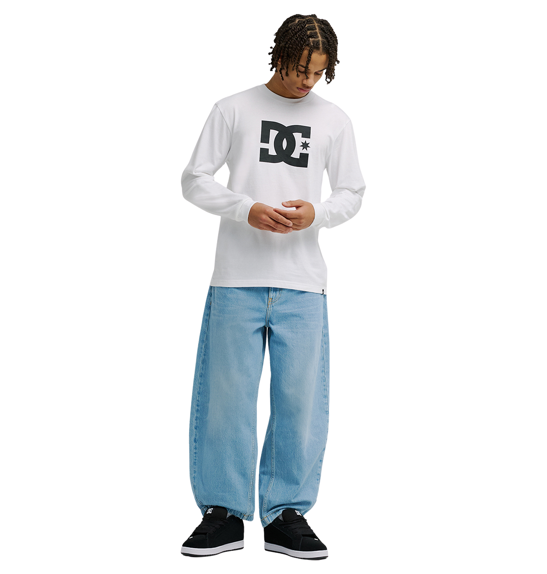 A person with short dreadlocks wears the Men's DC Star Tee by DC Shoes—a white long-sleeve shirt with bold black DC artwork—paired with loose light-blue jeans and black sneakers, standing against a plain white background looking down.