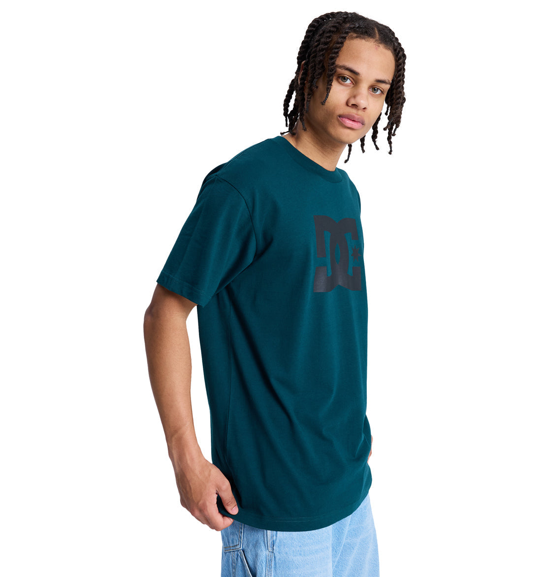 A young person with braided hair wears a teal Reebok DC Star Hss t-shirt and light blue jeans, standing against a plain white background and gazing slightly to the side.