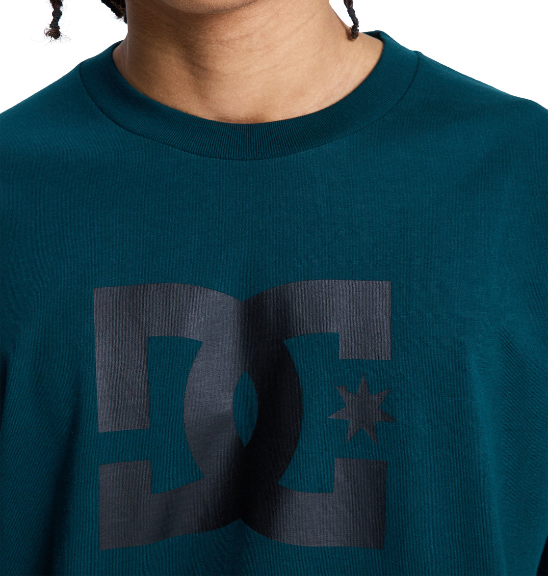 A person wears a teal Reebok DC Star Hss t-shirt featuring a large black DC logo with a star on the chest; only their lower face, neck, and upper torso are visible.