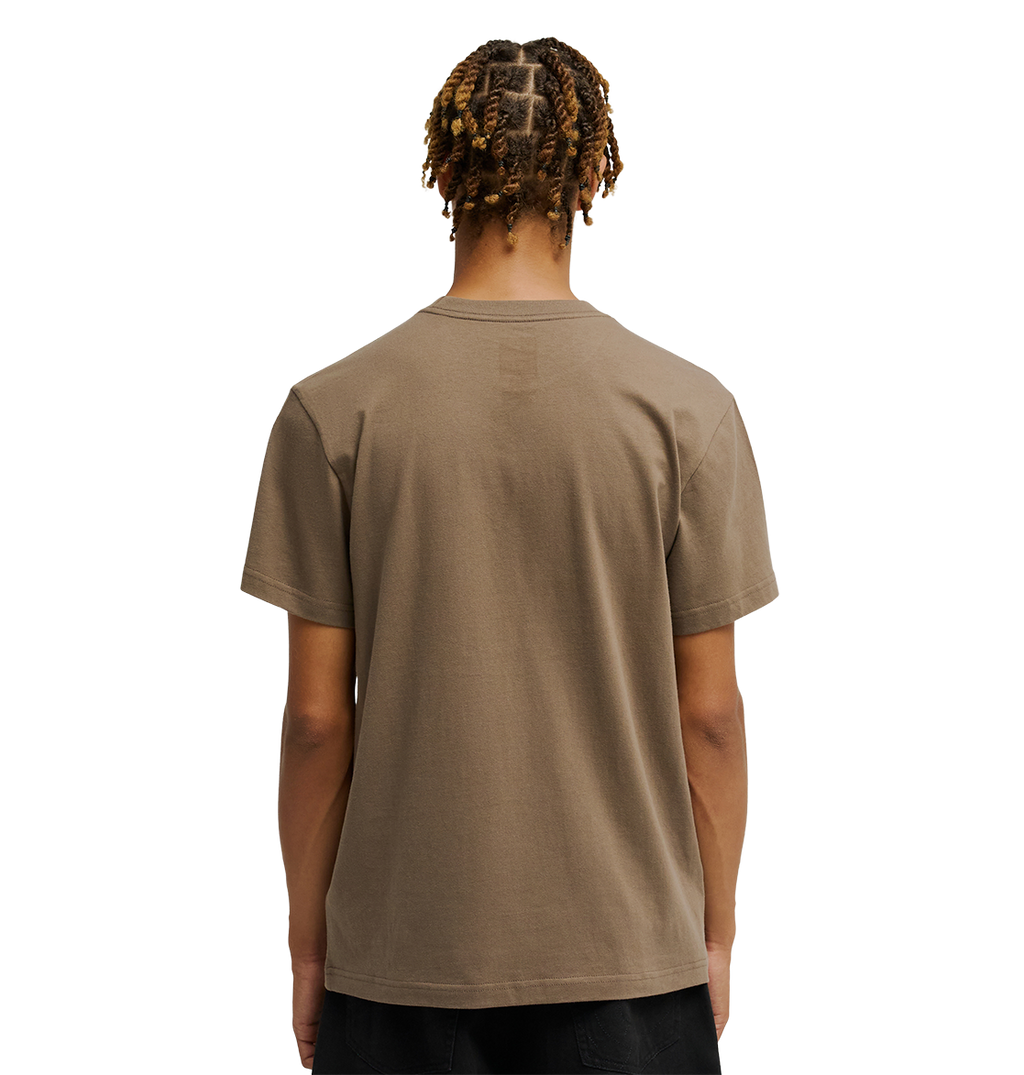 A person with short, twisted hair faces away, wearing the Men's DC Star Tee by DC Shoes in taupe and black pants against a white background.