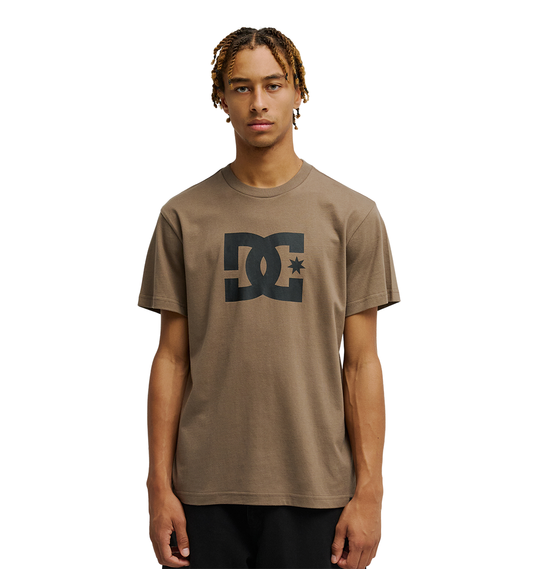A young man with short, curly blond hair wears the Men's DC Star Tee by DC Shoes, a relaxed fit brown shirt with bold black DC artwork on the chest. He stands facing forward against a plain white background.
