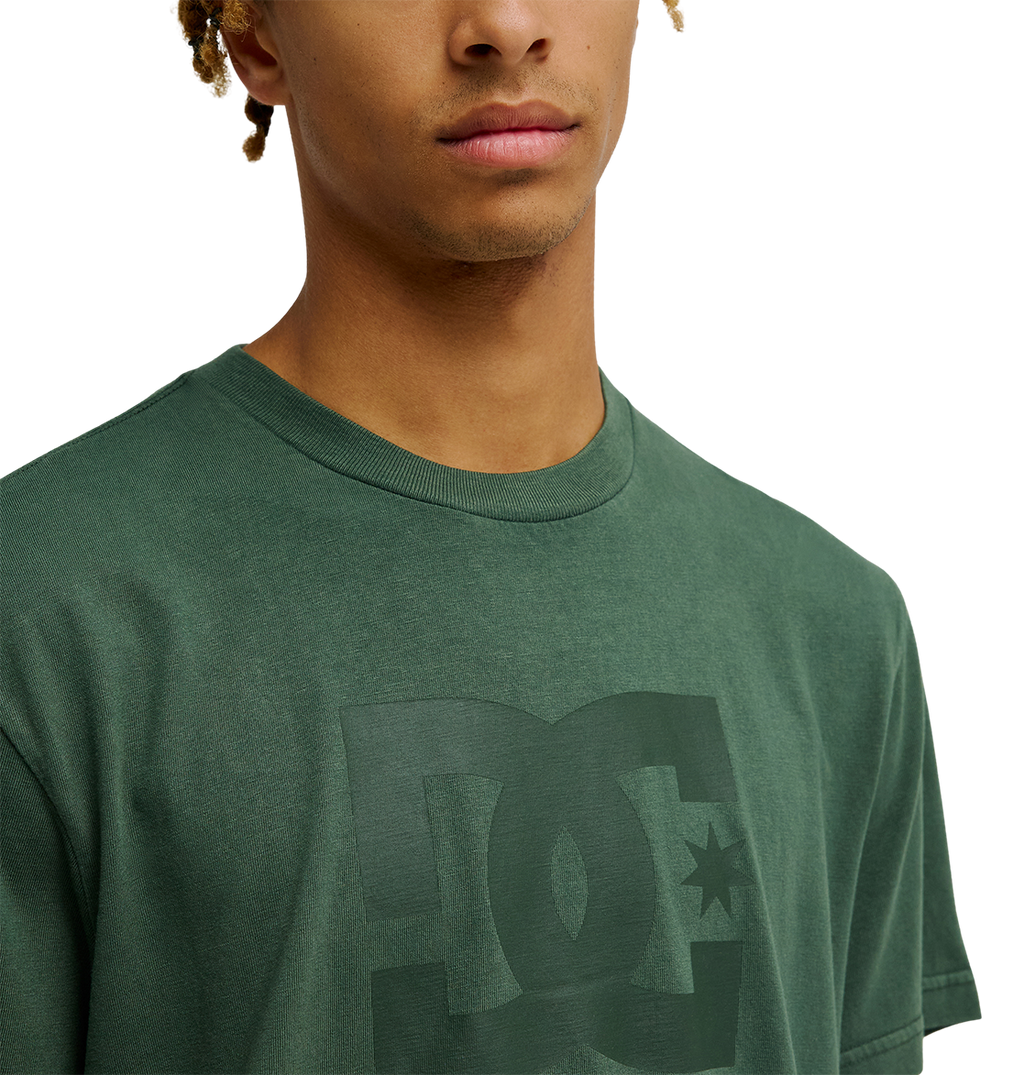 A person is shown from the shoulders up against a plain background, wearing the Men's DC Star Pigment Dye Tee by DC Shoes—a green skate t-shirt featuring a subtle logo on the front and a relaxed fit.