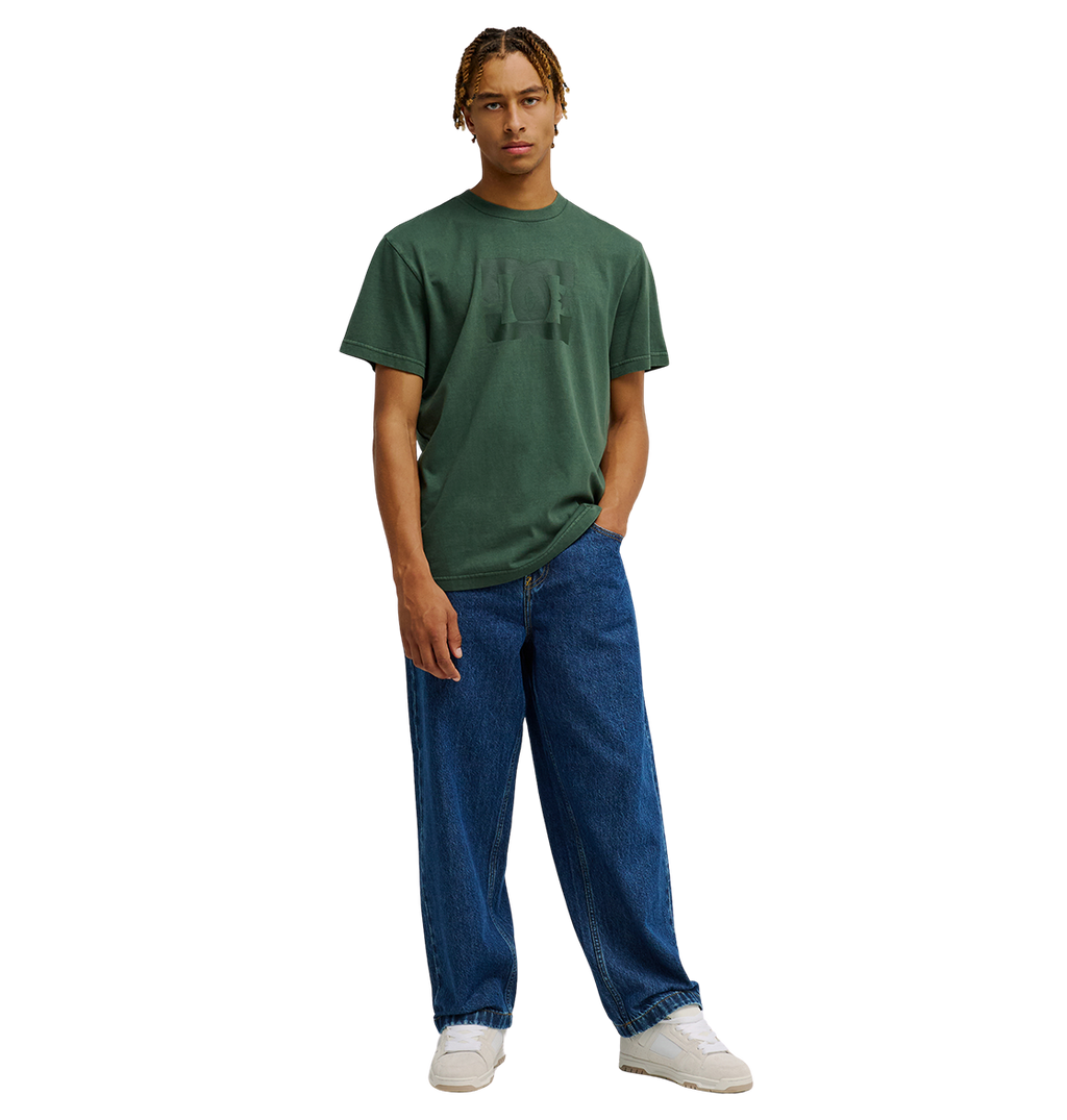 A person stands against a plain background wearing the Men's DC Star Pigment Dye Tee by DC Shoes in a relaxed fit dark green shade, paired with loose-fitting blue jeans and white sneakers, with one hand in their pocket.