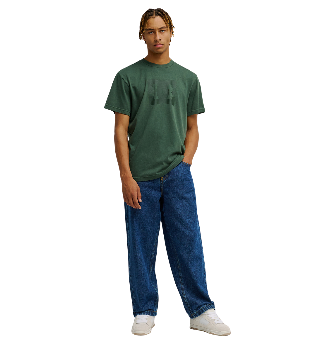 A person stands against a plain background wearing the Men's DC Star Pigment Dye Tee by DC Shoes in a relaxed fit dark green shade, paired with loose-fitting blue jeans and white sneakers, with one hand in their pocket.