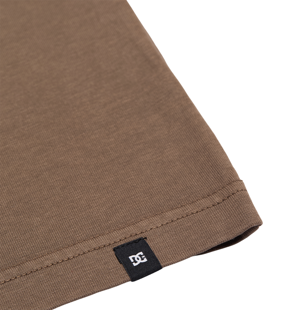 Close-up of the hem of the Men's DC Star Pigment Dye Tee by DC Shoes in brown, featuring a small black tag with a white DC logo sewn into the seam. The background is plain white.
