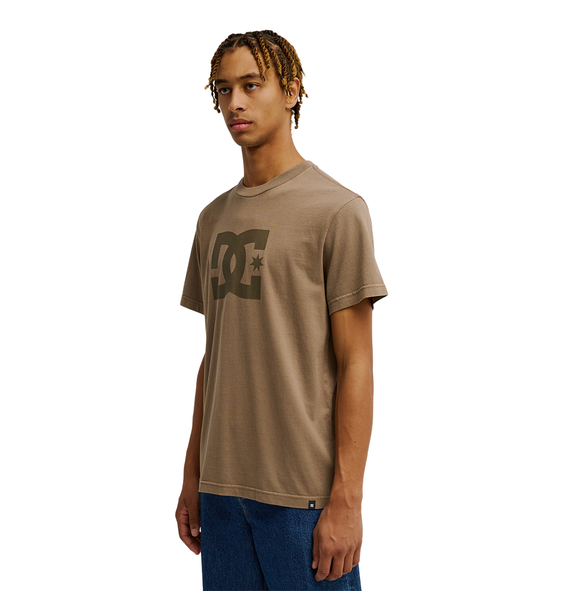 A young man with short, twisted hair wears the Men's DC Star Pigment Dye Tee by DC Shoes—a taupe graphic skate shirt featuring signature DC artwork—paired with blue jeans. He stands against a plain white background, gazing slightly to the side with a neutral expression.