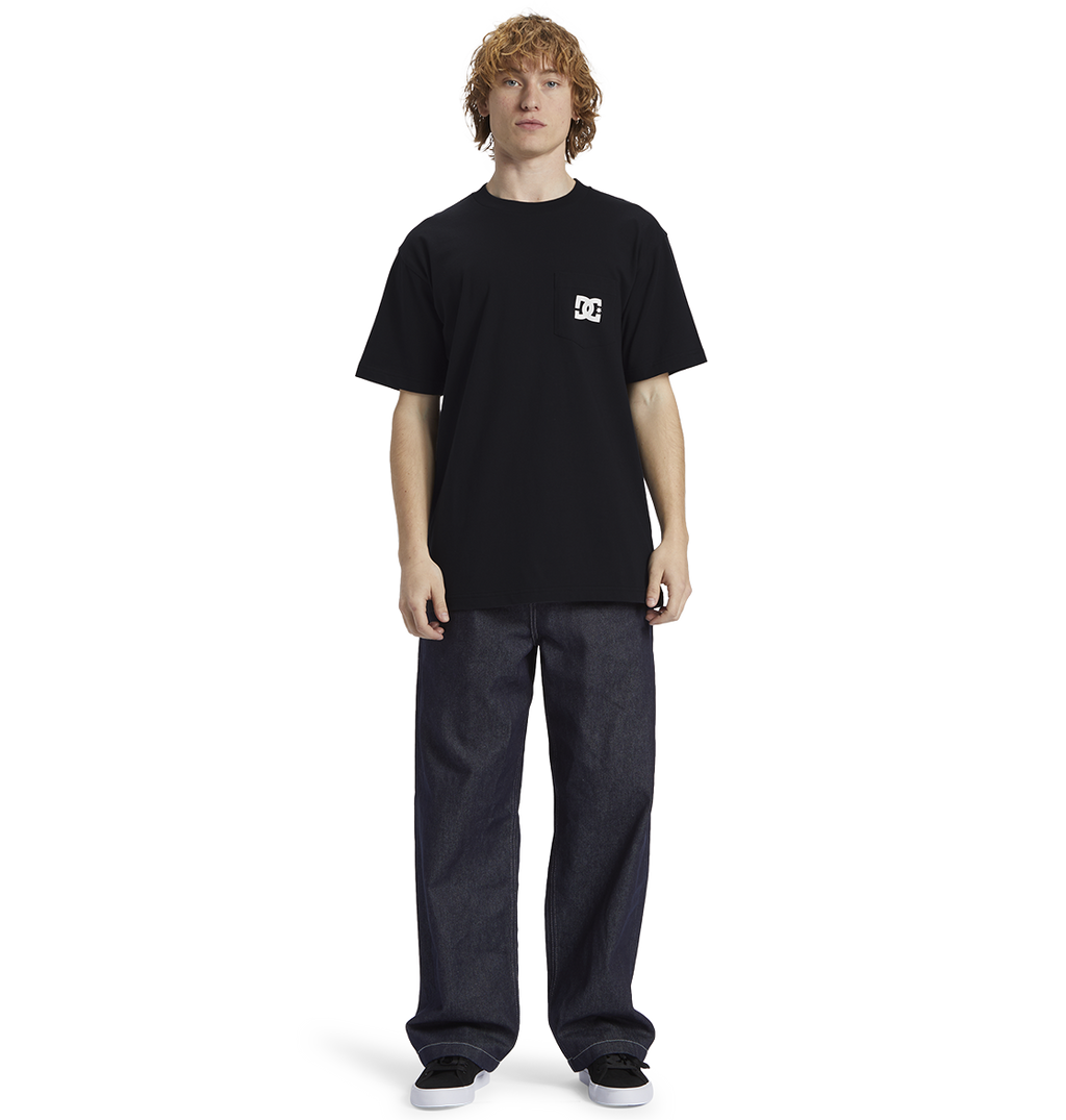 A person with curly hair wears the Men's DC Star Pocket Tee by DC Shoes in black, paired with relaxed fit dark jeans and black shoes, standing against a plain white background.