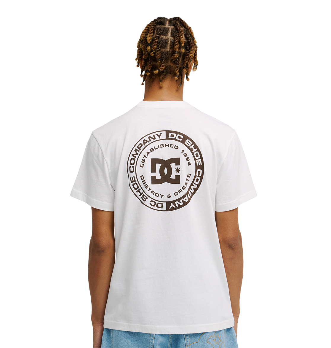 A person faces away wearing the Men’s DC Corpo Tee by DC Shoes, featuring a large circular skate-inspired logo and black text on the back, paired with light blue jeans. The background is plain white.