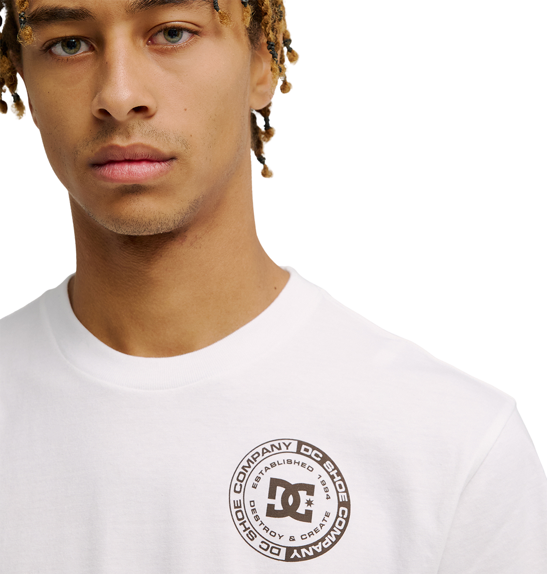 A young man wears the Men's DC Corpo Tee by DC Shoes, a relaxed fit white skate t-shirt with a circular corporate logo and surrounding text on the upper left chest. The background is plain white.