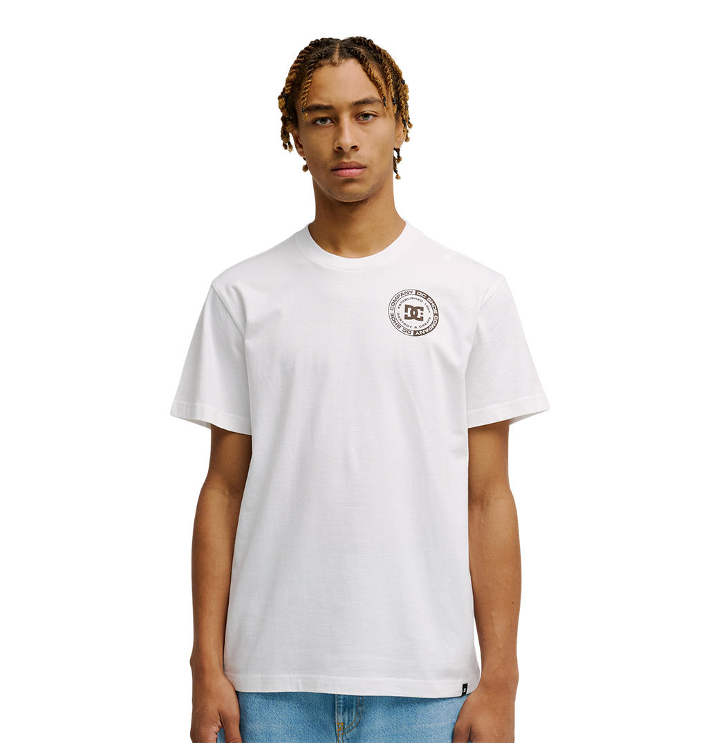 A young man with short, braided hair models the Men's DC Corpo Tee by DC Shoes—a relaxed fit, plain white t-shirt with a small black DC logo and circular emblem on the left chest—paired with light blue jeans against a white background.