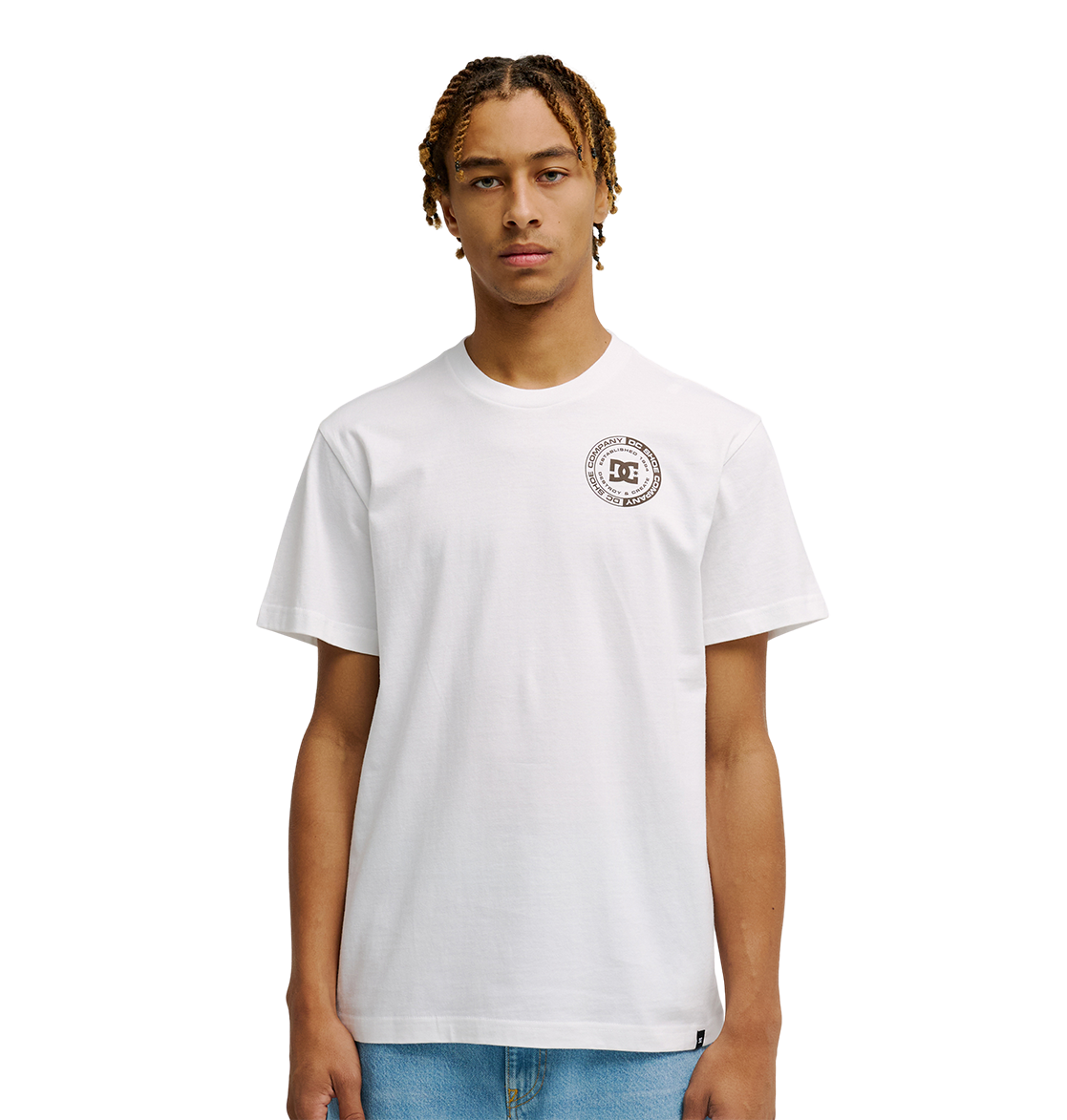 A young man with short, braided hair models the Men's DC Corpo Tee by DC Shoes—a relaxed fit, plain white t-shirt with a small black DC logo and circular emblem on the left chest—paired with light blue jeans against a white background.