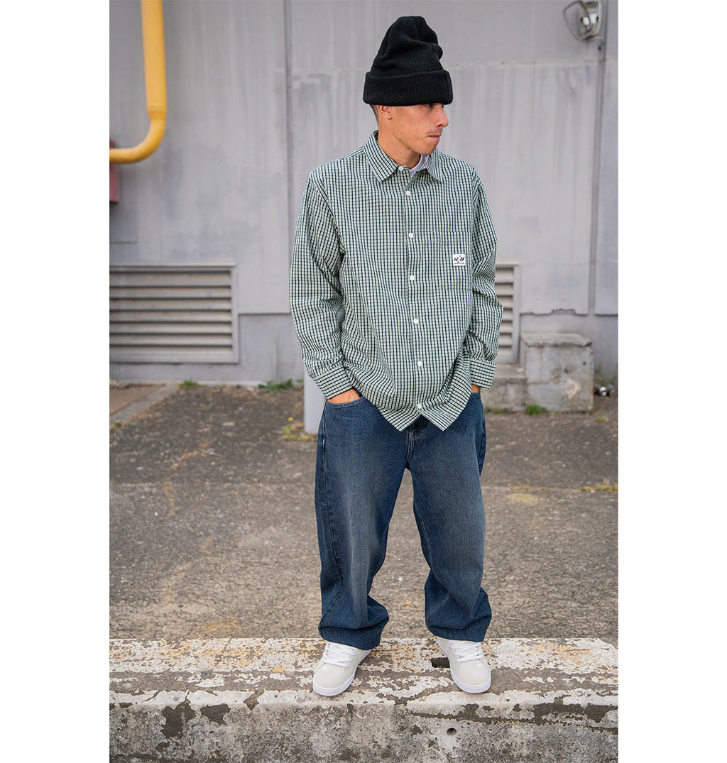 A man wearing a black beanie, green checkered button-up shirt, and white sneakers stands outdoors on pavement with his hands in his pockets, sporting classic 90s skate style in DC Shoes Men’s Worker Baggy Denim Double That Jeans.
