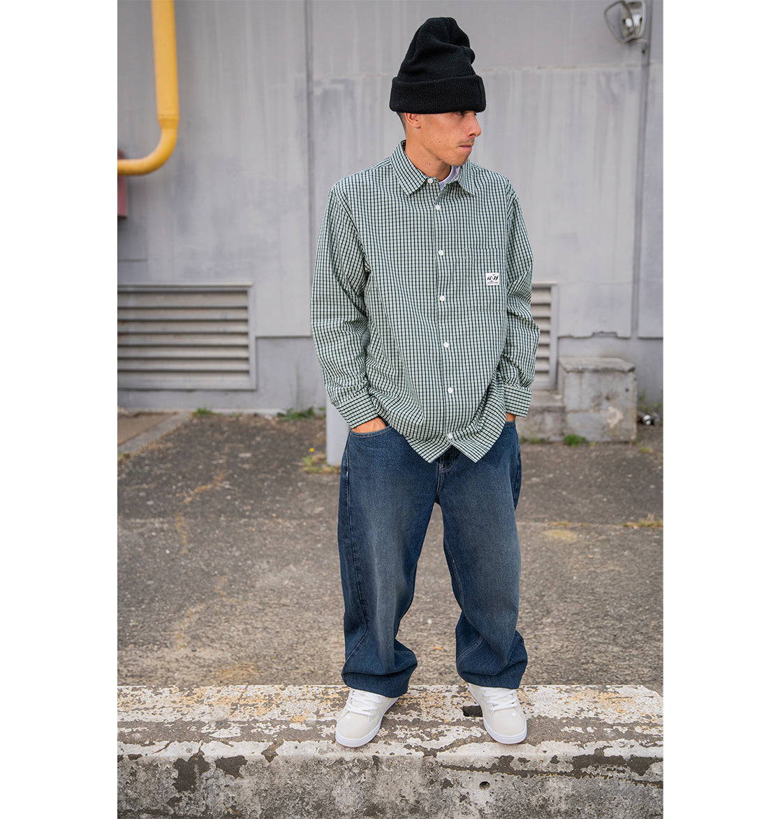 A man wearing a black beanie, green checkered button-up shirt, and white sneakers stands outdoors on pavement with his hands in his pockets, sporting classic 90s skate style in DC Shoes Men’s Worker Baggy Denim Double That Jeans.