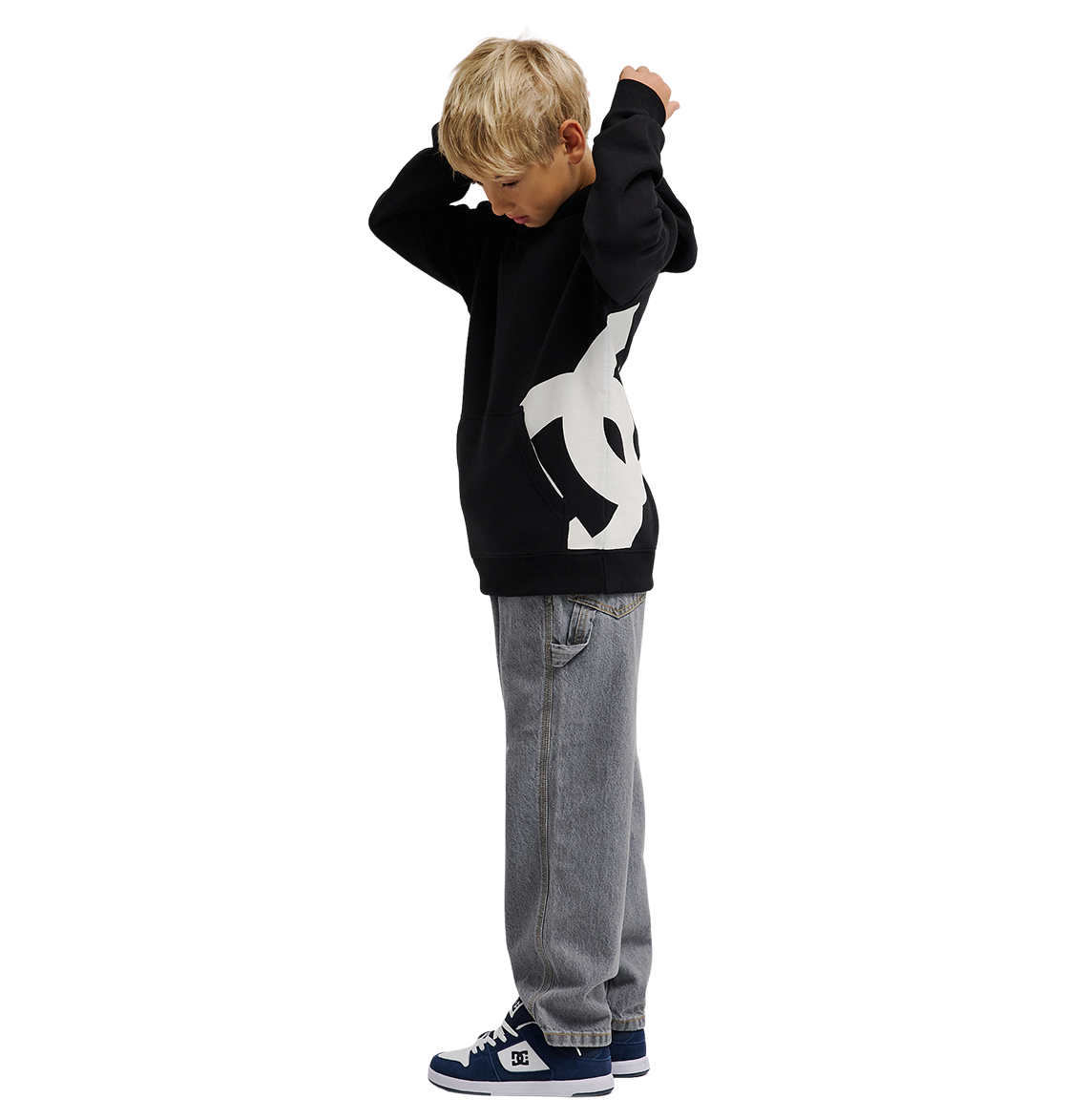 A young boy with blonde hair wears the DC Shoes Lanai Hoodie Youth—black with bold DC graphics—paired with gray jeans and blue sneakers, standing sideways and adjusting his hood for warmth against a white background.