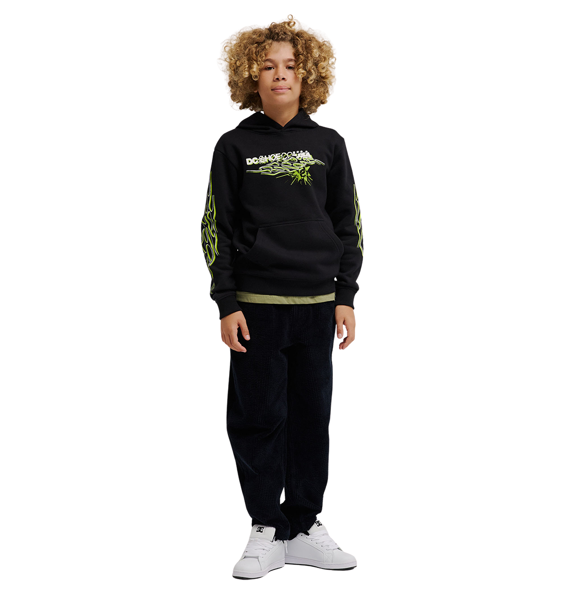 A child with curly hair wears the DC Shoes Side Flame Hoodie Youth, a black fleece hoodie with neon green DC graphics, paired with dark pants and white sneakers, standing facing forward against a plain white background.