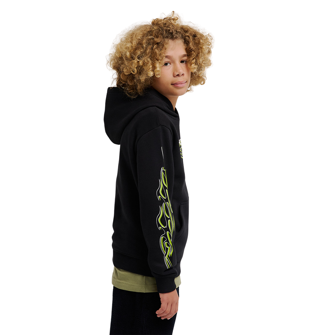 A youth model with curly blonde hair wears the DC Shoes Side Flame Hoodie, a black fleece featuring neon green flame designs on the sleeve, standing sideways and looking at the camera against a plain white background.