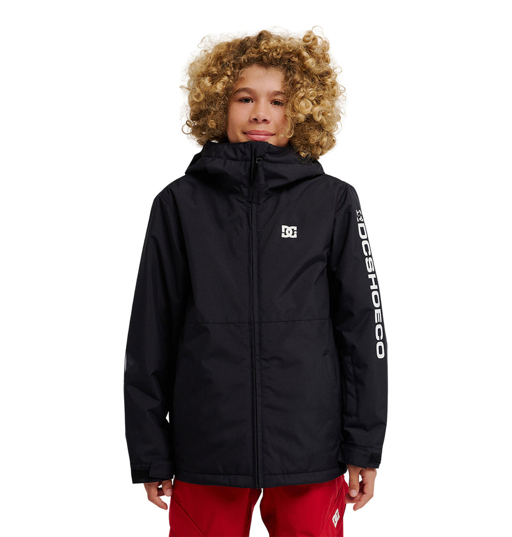 A young person with curly blond hair stands against a white background, wearing the DC Shoes Basis Youth Jacket—a black, durable, and stylish hooded jacket with DCSHOECO printed on the left sleeve—paired with red pants.