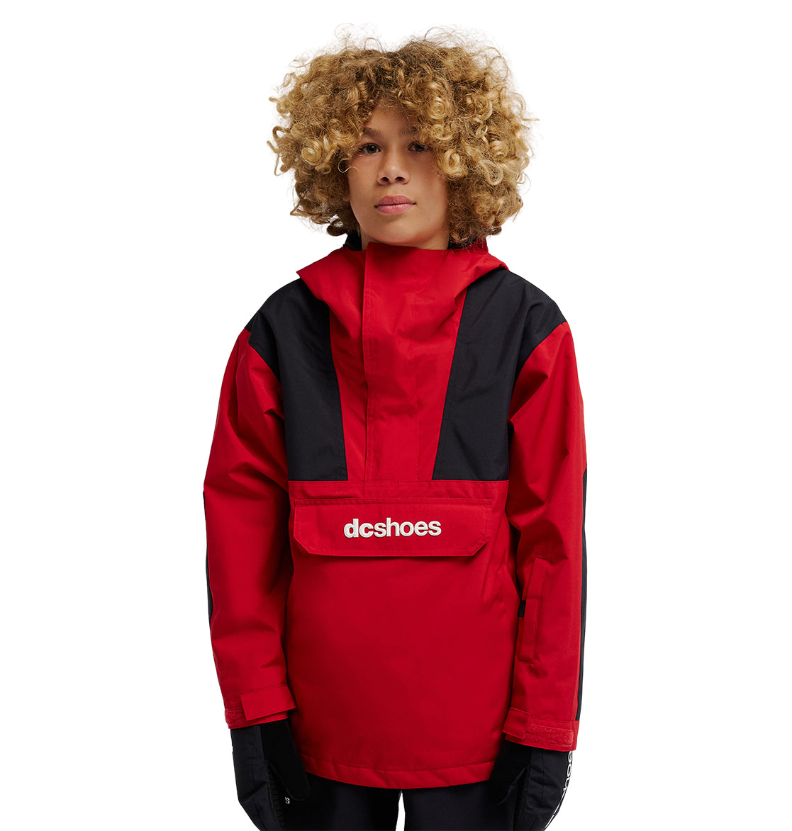 A person with curly blond hair wears the DC Shoes DC-43 Youth Anorak, a red and black insulated snow jacket featuring the “dcshoes” logo on the front pocket, standing against a plain white background.